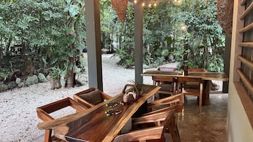 Un área de comedor exterior de madera con mesas y sillas, rodeada de exuberante vegetación y luces colgantes.