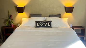 Ein ordentlich bezogenes Bett mit weißen Leinen, zwei Nachttischlampen, eine Zimmerpflanze auf dem Nachttisch und ein Kopfkissen mit der Aufschrift 'LOVE' und einem Herzdesign.