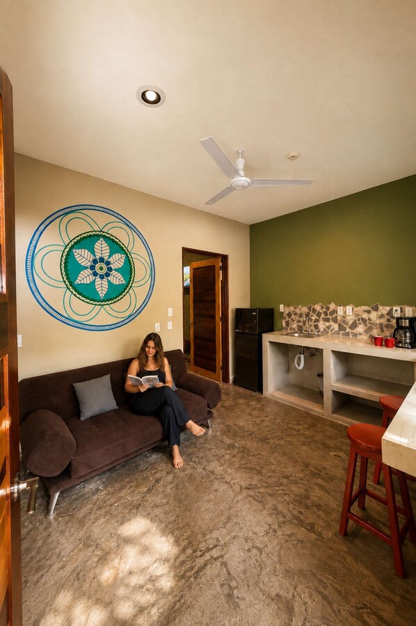 Una sala de estar con un sofá marrón, un ventilador de techo, un adorno en la pared, una zona de cocina con salpicadero de azulejos y un refrigerador.