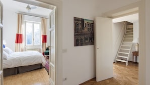 A bedroom with a bed, a window with curtains, a painting on the wall, and a staircase leading to another room. - Popolo Square Luxury Two Bedroom Apartment (Roma)
