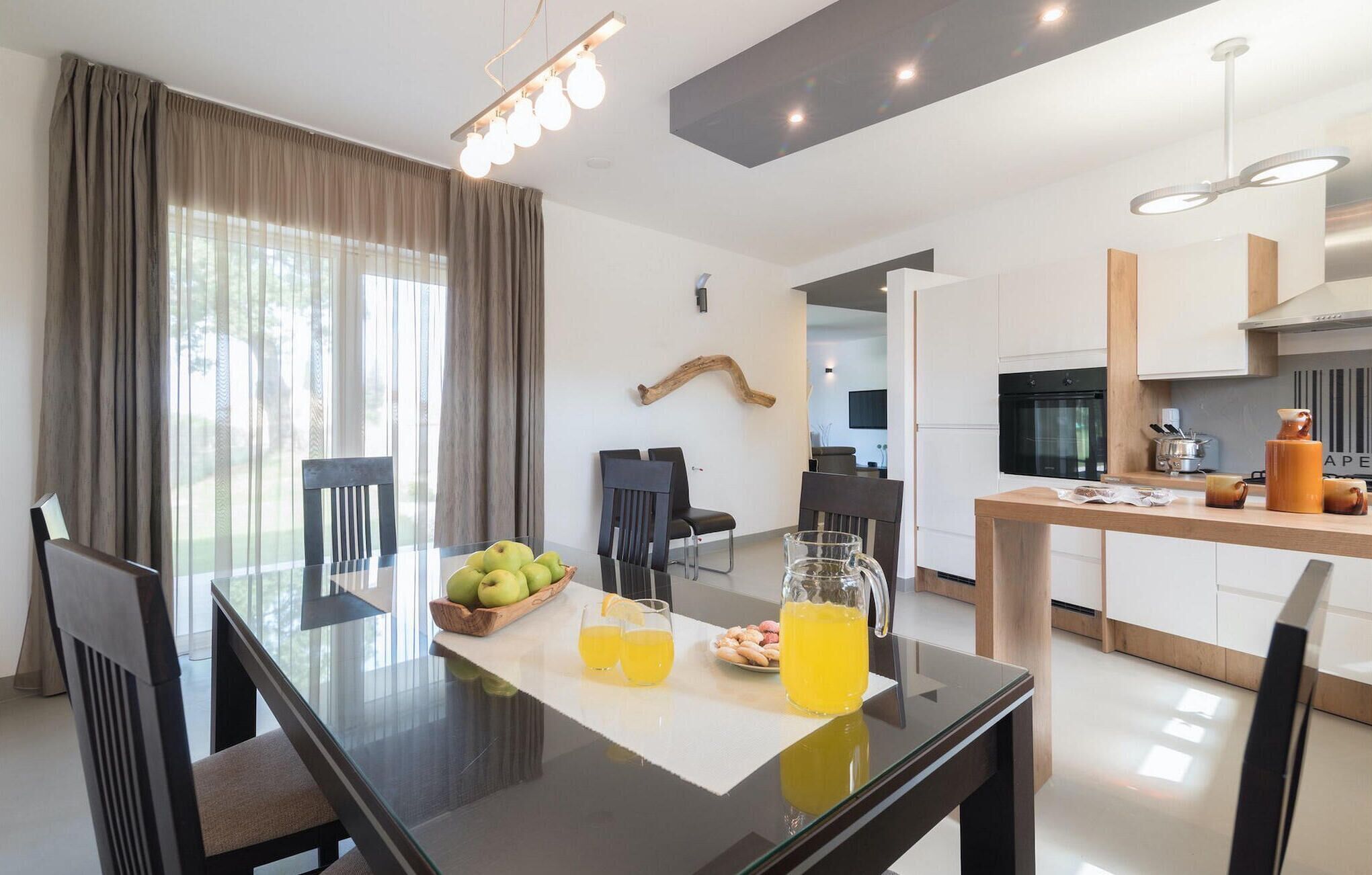 Un moderno comedor con una mesa de vidrio, sillas y un frutero. Se observa una cocina al fondo con armarios blancos y una estufa.