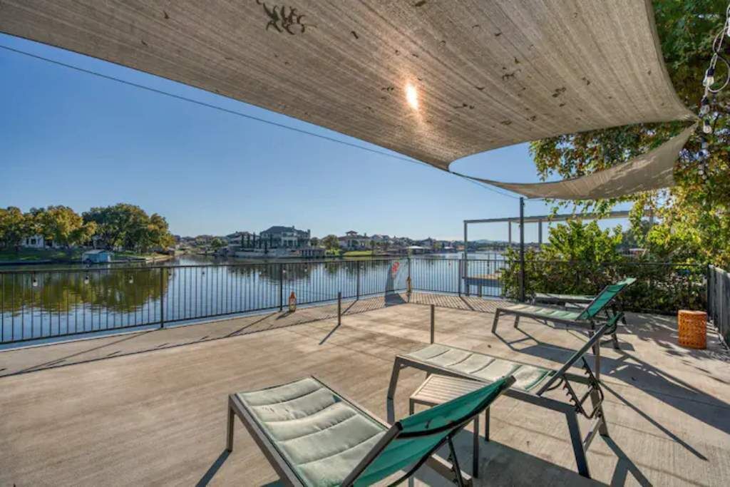 A terrace with sun loungers overlooking a lake, a concrete ceiling, and a view of buildings across the water.