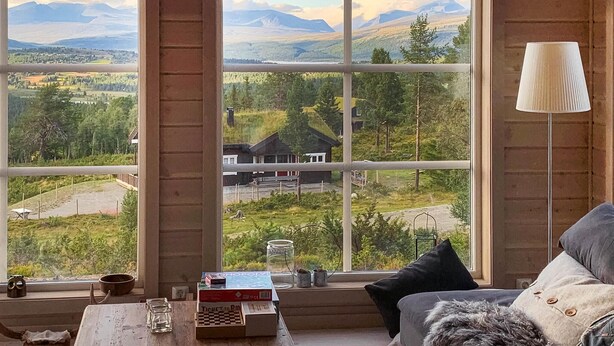 Ein Zimmer mit einem großen Fenster, das einen Blick auf eine gebirgige Landschaft freigibt. Im Raum steht ein Holztisch mit einem Schachbrett, einer Lampe und einigen Dekorationsgegenständen.