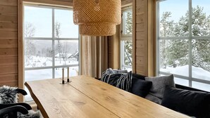 Ein Holzespeisetisch mit einer gewebten Pendelleuchte, eine Couch mit Kissen und große Fenster mit Blick auf eine verschneite Landschaft.