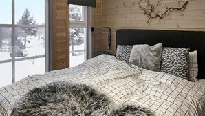 Ein Schlafzimmer mit einem großen Bett, einem hölzernen Kopfteil und Blick auf eine verschneite Landschaft durch ein Fenster.
