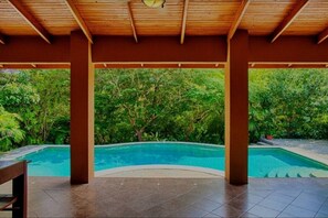 A covered outdoor swimming pool area with a wooden roof and tiled flooring, surrounded by lush greenery. - Henton Hacienda ,fiv minutes drive from the beach. (Los Pargos)