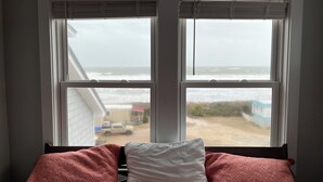 Une chambre avec un lit adossé à un mur, deux fenêtres équipées de stores, et une vue sur la plage et l’océan.