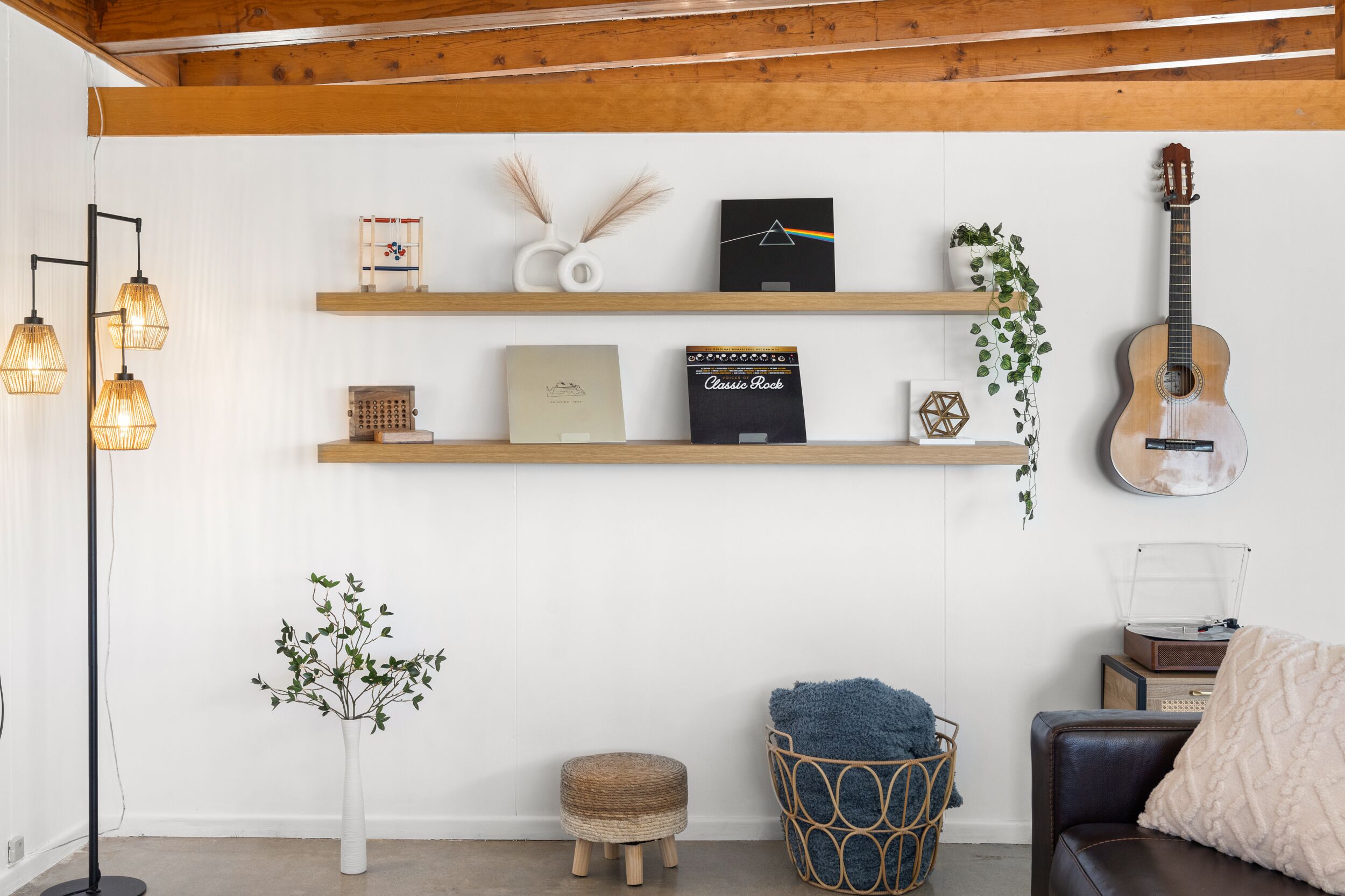 A room with wooden beams, a guitar, shelves with books and decorative items, a floor lamp, a small plant, and a cozy seating area.