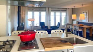 Una cocina moderna con estufa a gas, una olla roja y una tabla de cortar de madera. Hay un comedor con sillas y una mesa, y una ventana con cortinas.