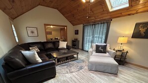 A cozy living room with a black leather sofa, a striped armchair, and a coffee table. A wooden ceiling and a large window with curtains are visible. - The Winterbrook Mountain House.
Bike Week Garage Owls Nest Golf (Campton)