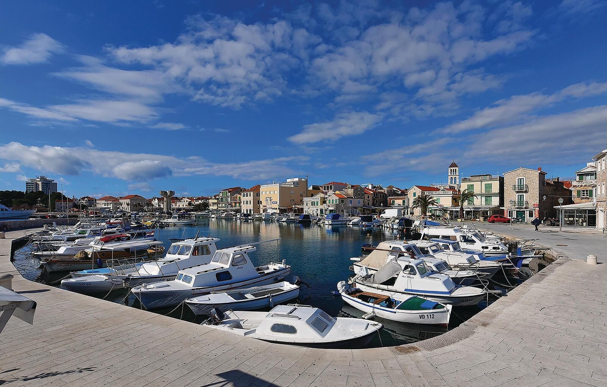 A marina with various boats docked, a clear sky, and buildings in the background.