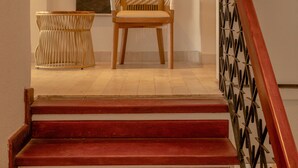 A staircase with red steps and a wooden handrail, leading to a room with a large framed picture of a building, a wicker chair, and a woven side table. - Casa Griega By Wynwood House (Cartagena)
