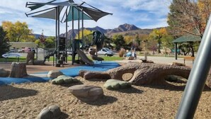 Un’area giochi con scivolo e strutture per arrampicata, circondata da rocce e alberi, con montagne sullo sfondo.