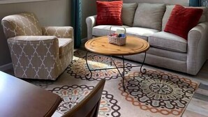 Una sala de estar acogedora con ventilador de techo, un sofá con cojines rojos, un sillón estampado y una mesa de centro.