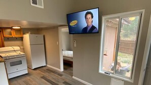 Cocina compacta con horno empotrado, refrigerador y estufa. Un televisor está montado en la pared, y hay una ventana con vista al patio trasero.