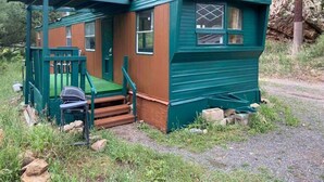 Een klein, gelijkvloers blokhutje met een veranda en een klein terras, omringd door natuurlijke begroeiing en een rotsachtige helling.