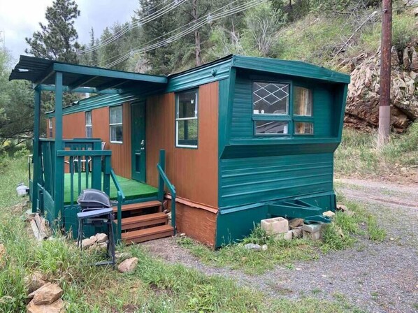 A small, single-story cabin with a porch and a small deck, surrounded by natural vegetation and a rocky hillside.