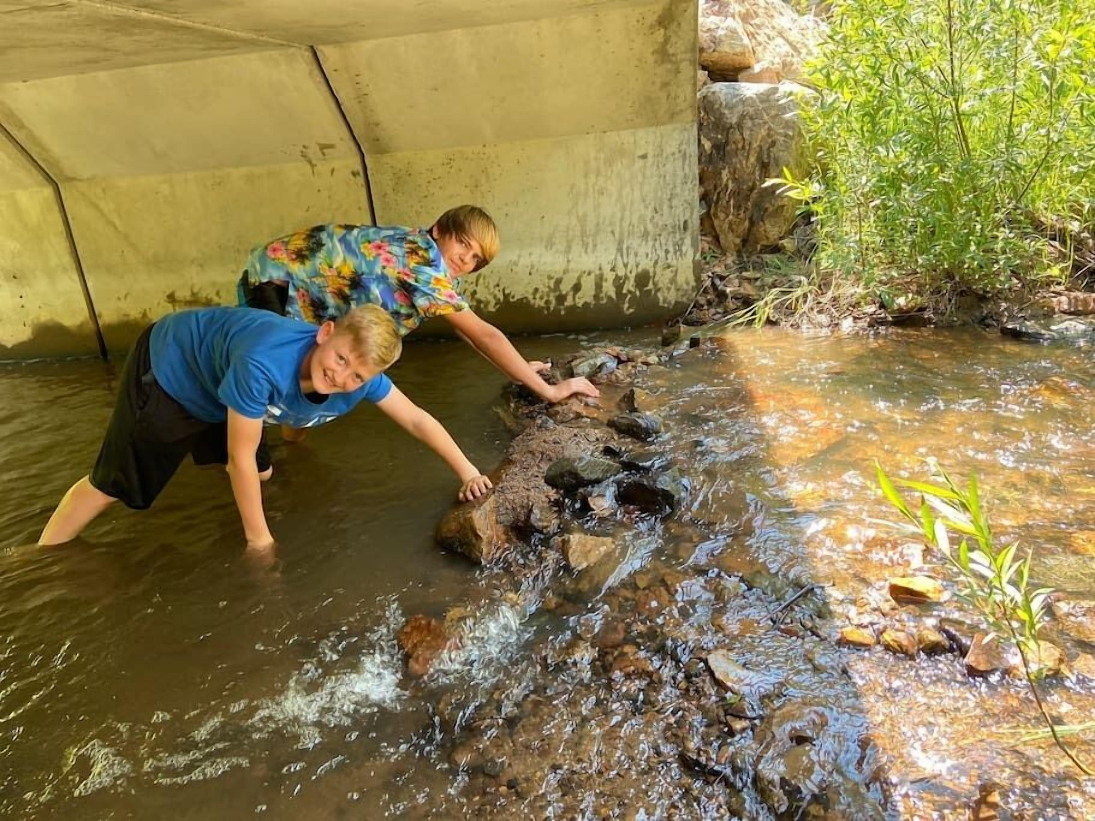 Two individuals crossing a shallow stream under a concrete bridge.