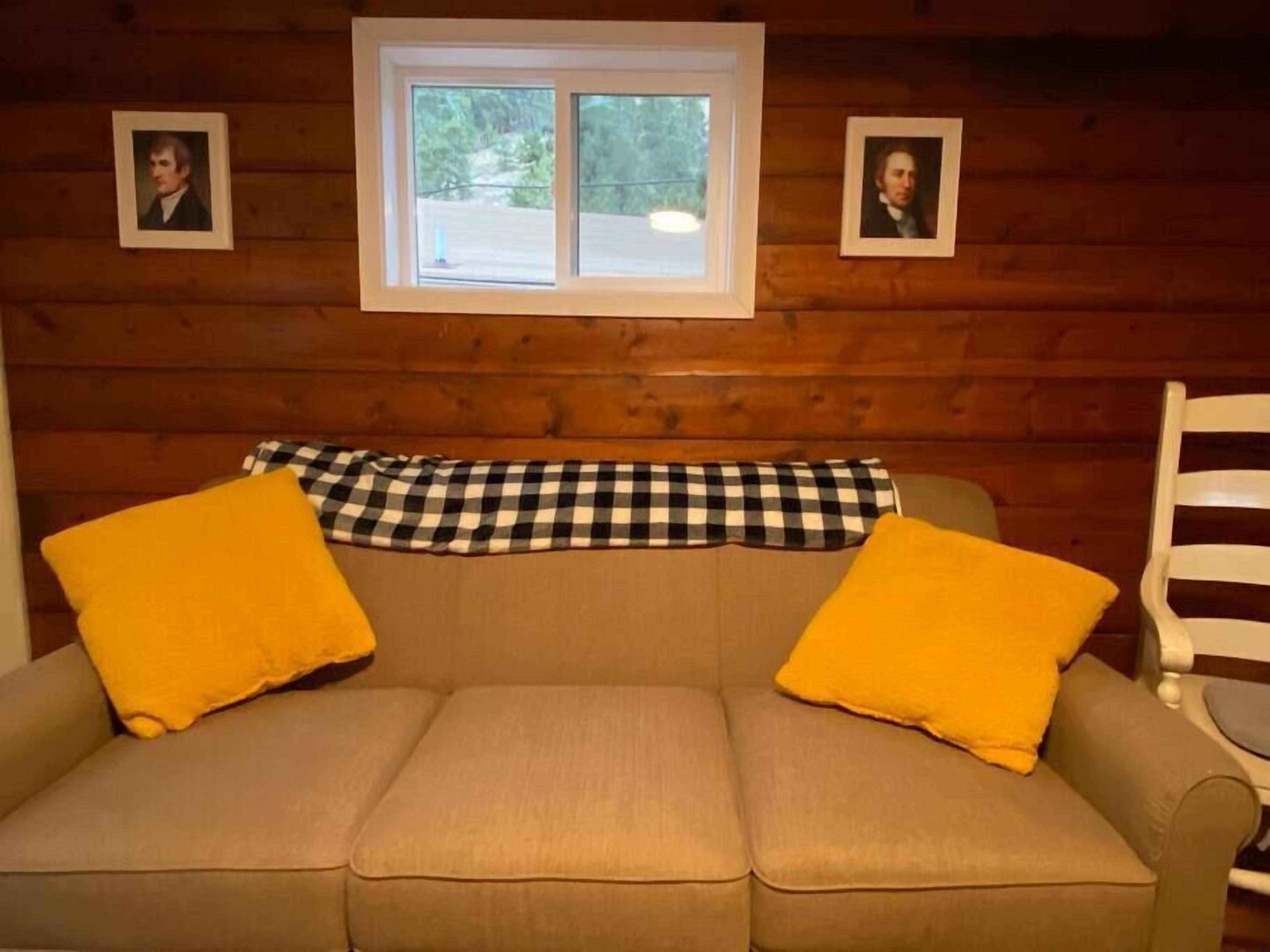 A wooden cabin interior with a beige sofa, two yellow pillows, a checkered throw, and two framed pictures on the wall.