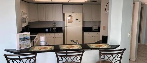 A compact kitchen with white cabinets, a refrigerator, and a microwave. There are three bar stools with decorative backs.