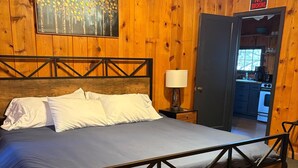 A wooden-paneled bedroom with a bed, pillows, a nightstand, a lamp, and a door leading to a kitchen. - Piedra River Outpost (Bayfield)