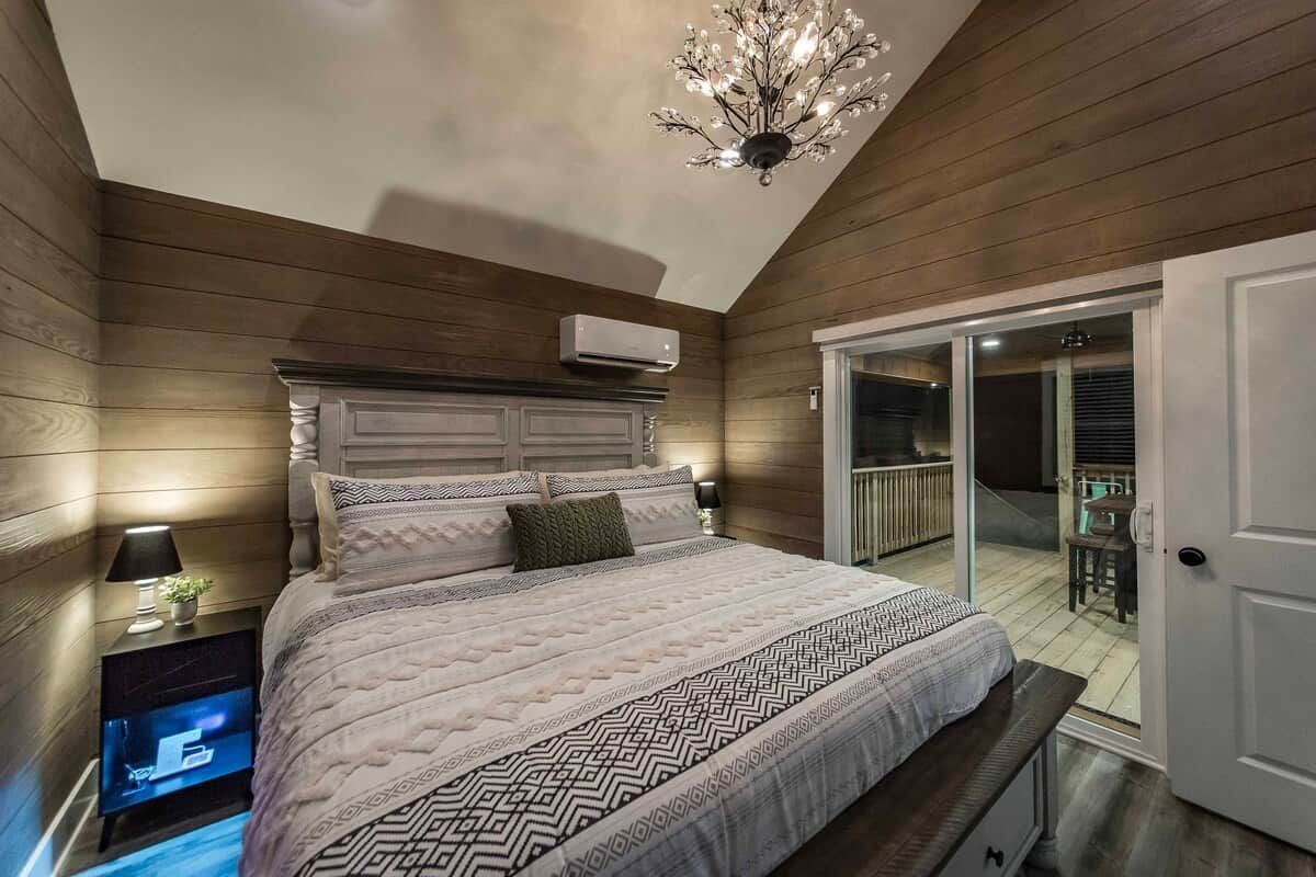 A bedroom with a large bed, a television on a stand, a chandelier, and a sliding glass door leading to an outdoor area.