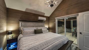 A bedroom with a large bed, a television on a stand, a chandelier, and a sliding glass door leading to an outdoor area.