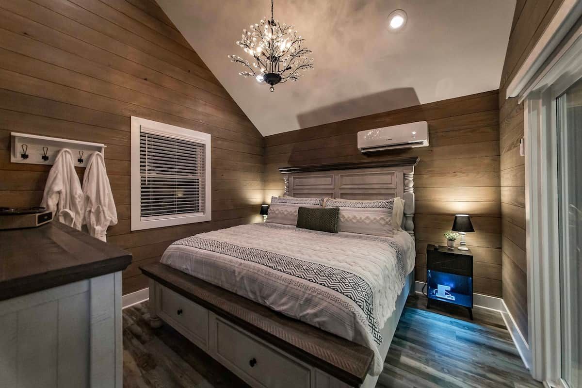 A bedroom with a wooden ceiling, a chandelier, a bed with patterned bedding, a nightstand with a lamp, and an air conditioning unit.