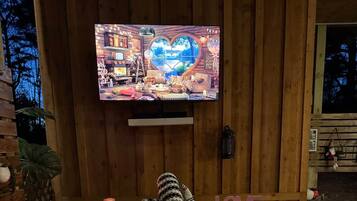 Interior de una cabaña de madera con televisión montada en la pared, ventana con forma de corazón y un jacuzzi con luz roja.