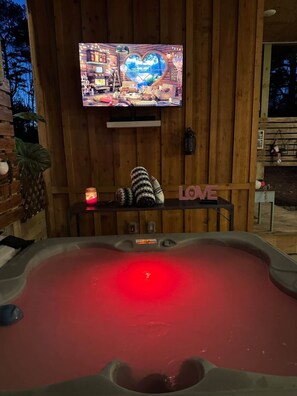 A wooden cabin interior with a television mounted on the wall, a heart-shaped window, and a hot tub with a red light.