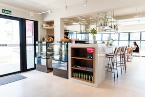 Interior moderno de una cafetería con un expositor de vidrio, estanterías de madera y un área de estar con sillas y mesas.