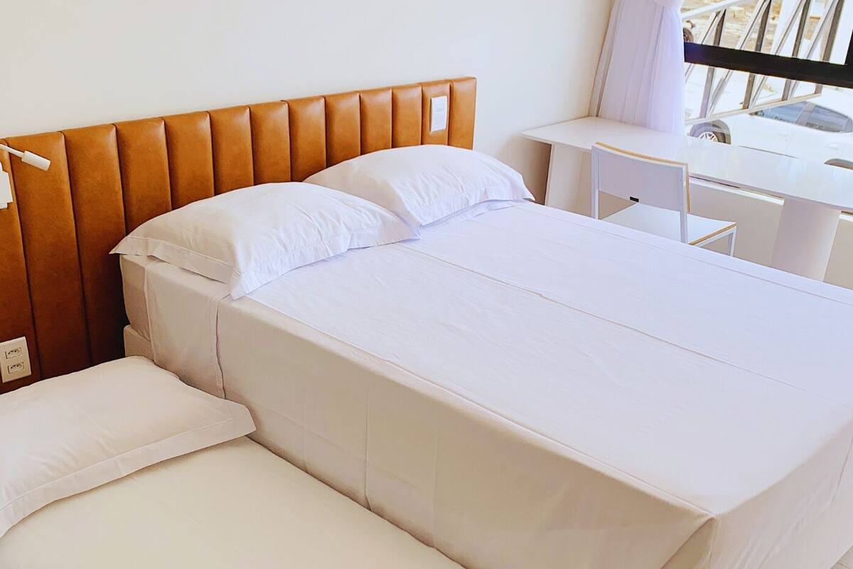 Habitación de hotel con cama, cabecera de madera, mesita de noche y ventana que da a la vista de un vehículo en el exterior.