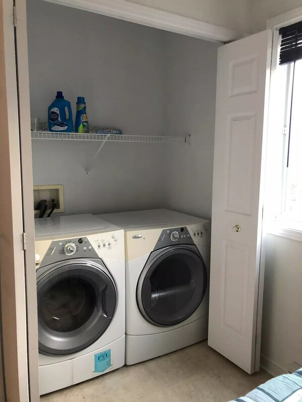 A laundry room with two front-loading washing machines, a shelf with detergent bottles, and a door to the right.