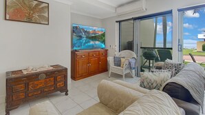 A spacious living room with a large TV, a wooden cabinet, a white sofa, and a view of the outdoors through a sliding glass door. - Magnetic Docks Townhouse 8 (Nelly Bay)
