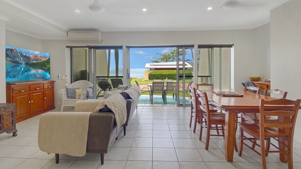 A spacious living area with a sofa, dining table, and chairs, a television, and a view of the outdoors. - Magnetic Docks Townhouse 8 (Nelly Bay)