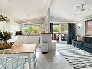 A modern kitchen with white cabinets, a dining area with a wooden table and chairs, a small trash bin, and a view of the outdoors through a sliding door.