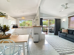 A modern kitchen with white cabinets, a dining area with a wooden table and chairs, a small trash bin, and a view of the outdoors through a sliding door. - Magnetic Haven Unit 7 (Arcadia)