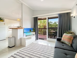 A modern living room with a flat-screen TV, a sofa, a dining area, and a balcony with a view of palm trees. - Magnetic Haven Unit 7 (Arcadia)