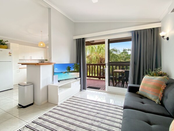 A modern living room with a flat-screen TV, a sofa, a dining area, and a balcony with a view of palm trees.