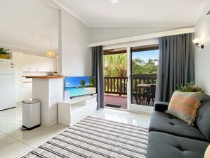 A modern living room with a flat-screen TV, a sofa, a dining area, and a balcony with a view of palm trees.