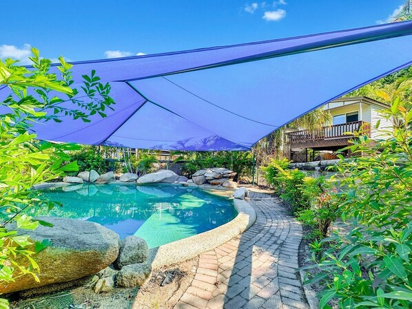 A swimming pool with a curved edge, surrounded by rocks and plants, under a blue canopy.