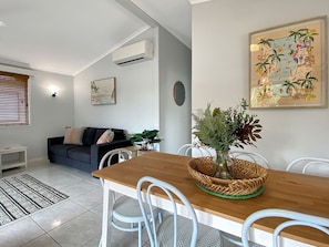 A modern living room with a wooden dining table, white chairs, a dark sofa, and a potted plant. - Magnetic Haven Unit 7 (Arcadia)