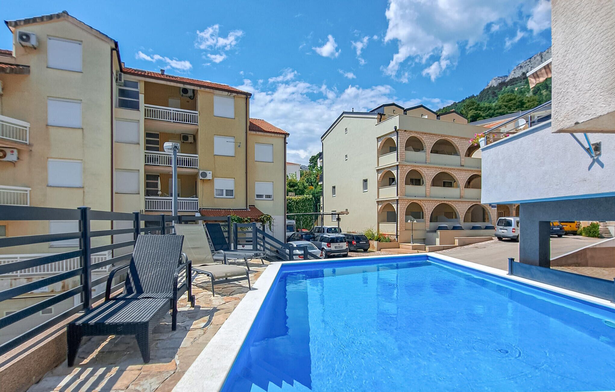 A swimming pool area with a bench and lounge chairs, surrounded by multi-story buildings with balconies and a mountain backdrop.