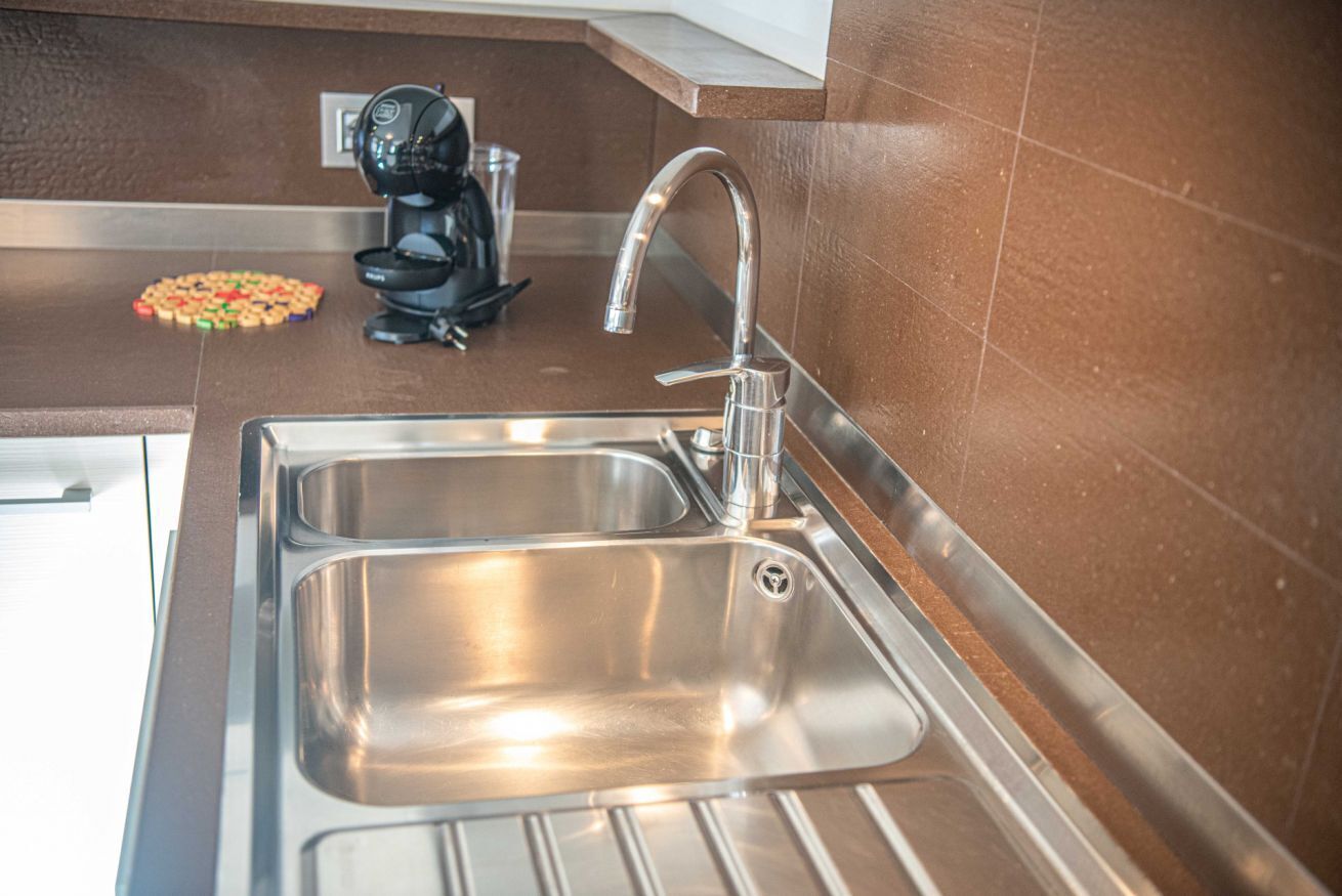 Un lavamanos de acero inoxidable con un grifo moderno, una cafetera y un recipiente de vidrio sobre la encimera.