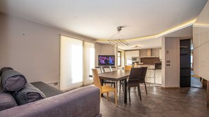 A modern living room with a sofa, a dining table with chairs, a TV, and a kitchen area in the background. - Convenience, comfort and atmosphere at your fingertips. (Livigno)
