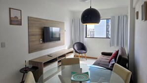 Een moderne woonkamer met een flatscreen televisie bevestigd aan een houten paneel, een glazen eettafel met stoelen, een zwart gestoffeerde bank en een groot raam met gordijnen.
