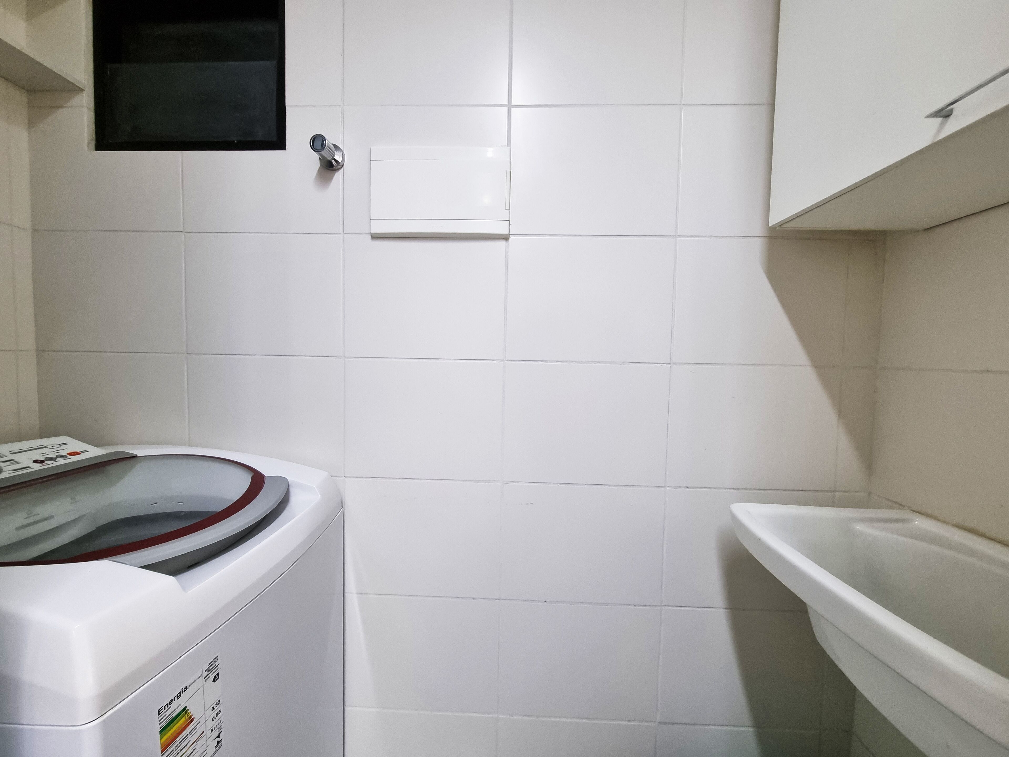 A laundry area with a washing machine, a white tiled wall, a small black shelf, and a white sink.