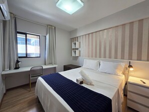 Une chambre d’hôtel comprenant un lit, un bureau, une chaise, une fenêtre avec des rideaux, une lampe de chevet et une étagère fixée au mur.