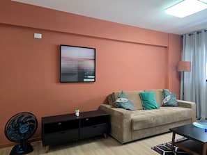 A living room with a sofa, a flat-screen TV mounted on the wall, a standing fan, and a lamp. - Apê a 3 min do Mar e Piscina na Cobertura (Maceió)