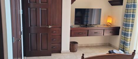 Una habitación con puerta de madera, televisor sobre un mueble de madera, una cesta de mimbre y una silla.